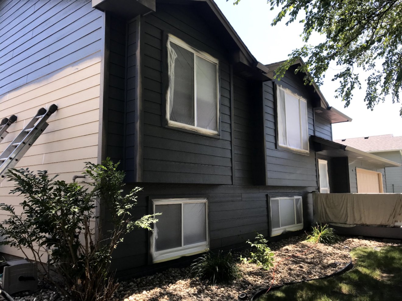 Exterior repaint in progress on a Sioux Falls home — siding freshly painted charcoal, windows masked off, ladder set against the wall
