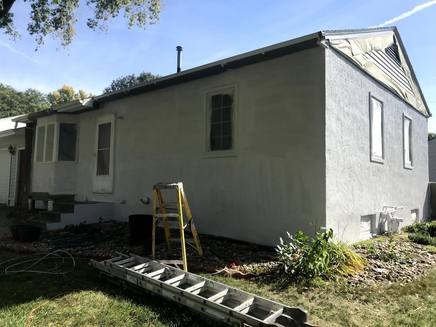 Single-story home exterior being repainted, ladder and equipment set up in the yard
