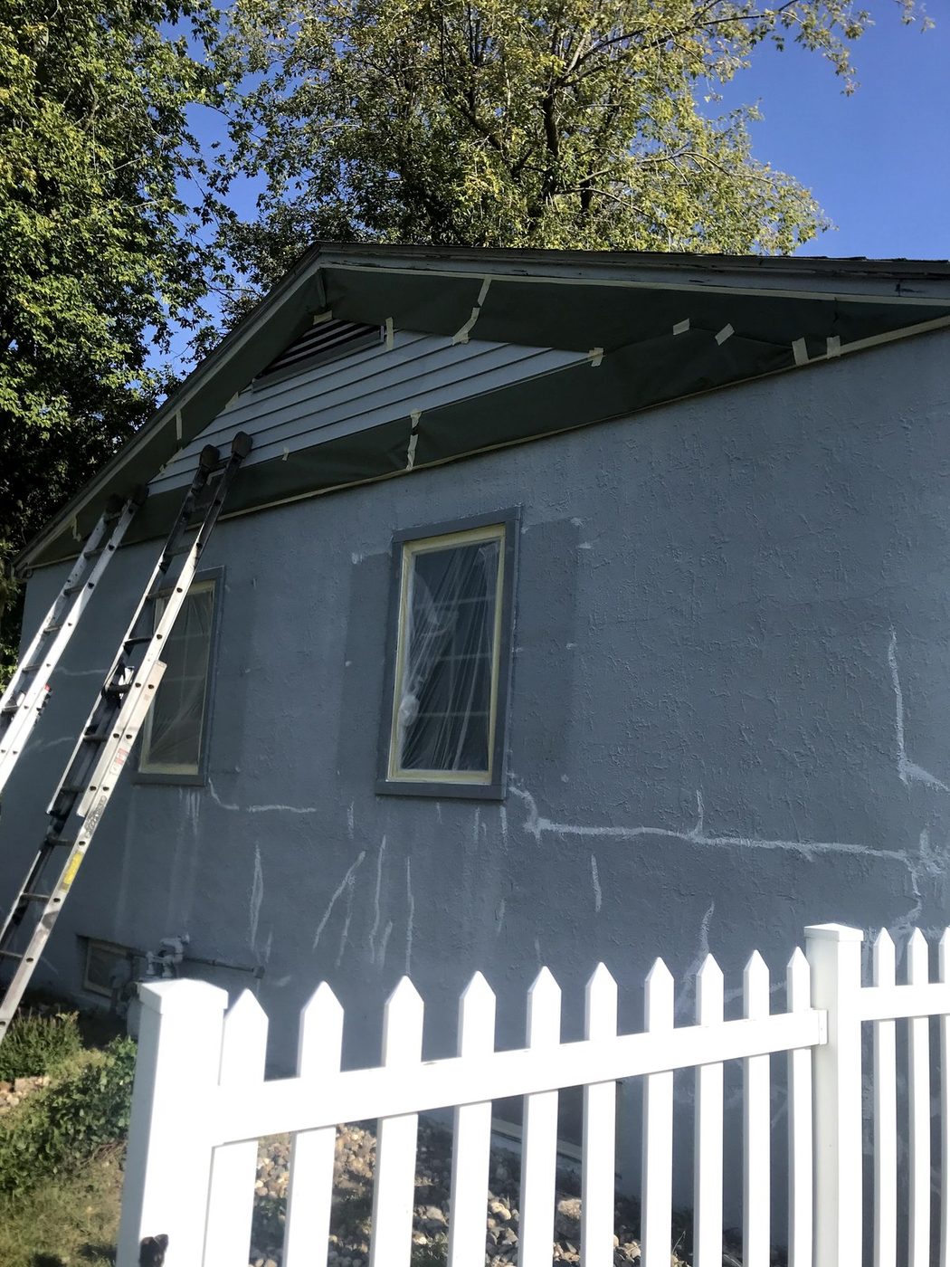Ladder against gray-blue siding mid-repaint, white picket fence in the foreground